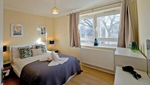 A hotel room with a bed, bedside table, lamp, window with curtains, radiator, and a view of trees outside. - Little Venice in Paddington central London 3 bedroom apartment (London)