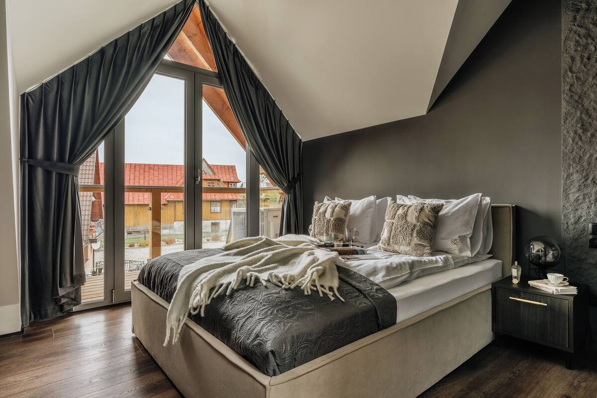 Un dormitorio con una cama grande, una mesita de noche y vistas a edificios a través de una ventana con cortinas.