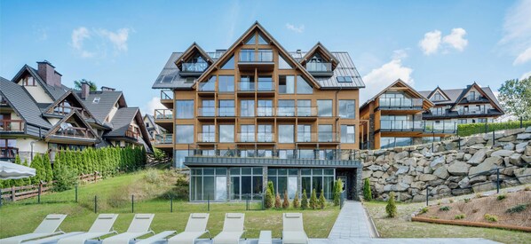 A modern multi-story building with a prominent central section, large windows, and a stone retaining wall. - Landscape Cyrhla with SPA by BQQK (Zakopane)