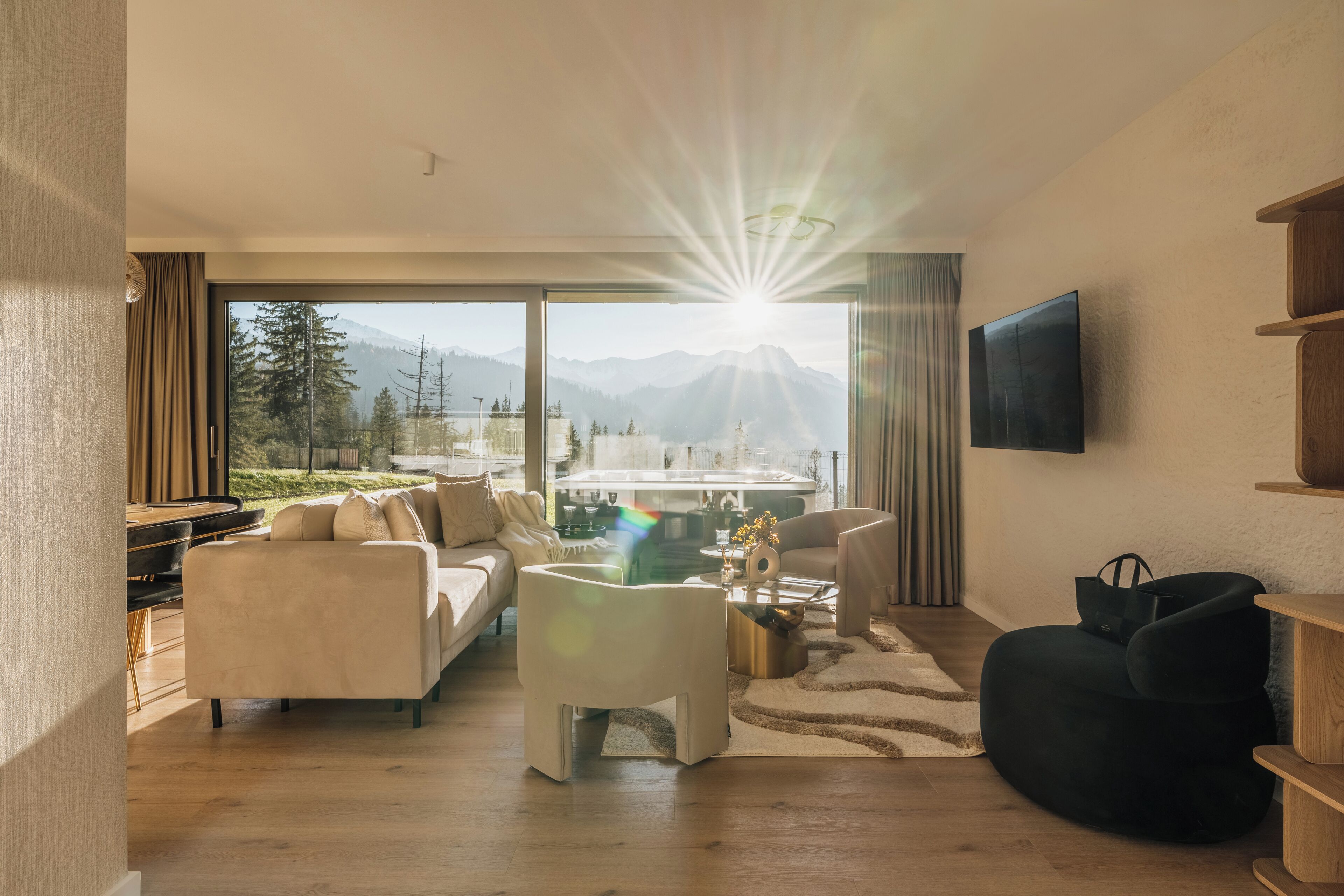 Uma sala de estar moderna com sofá, mesa de centro e banqueta, uma TV fixada na parede e uma grande janela com vista para as montanhas.