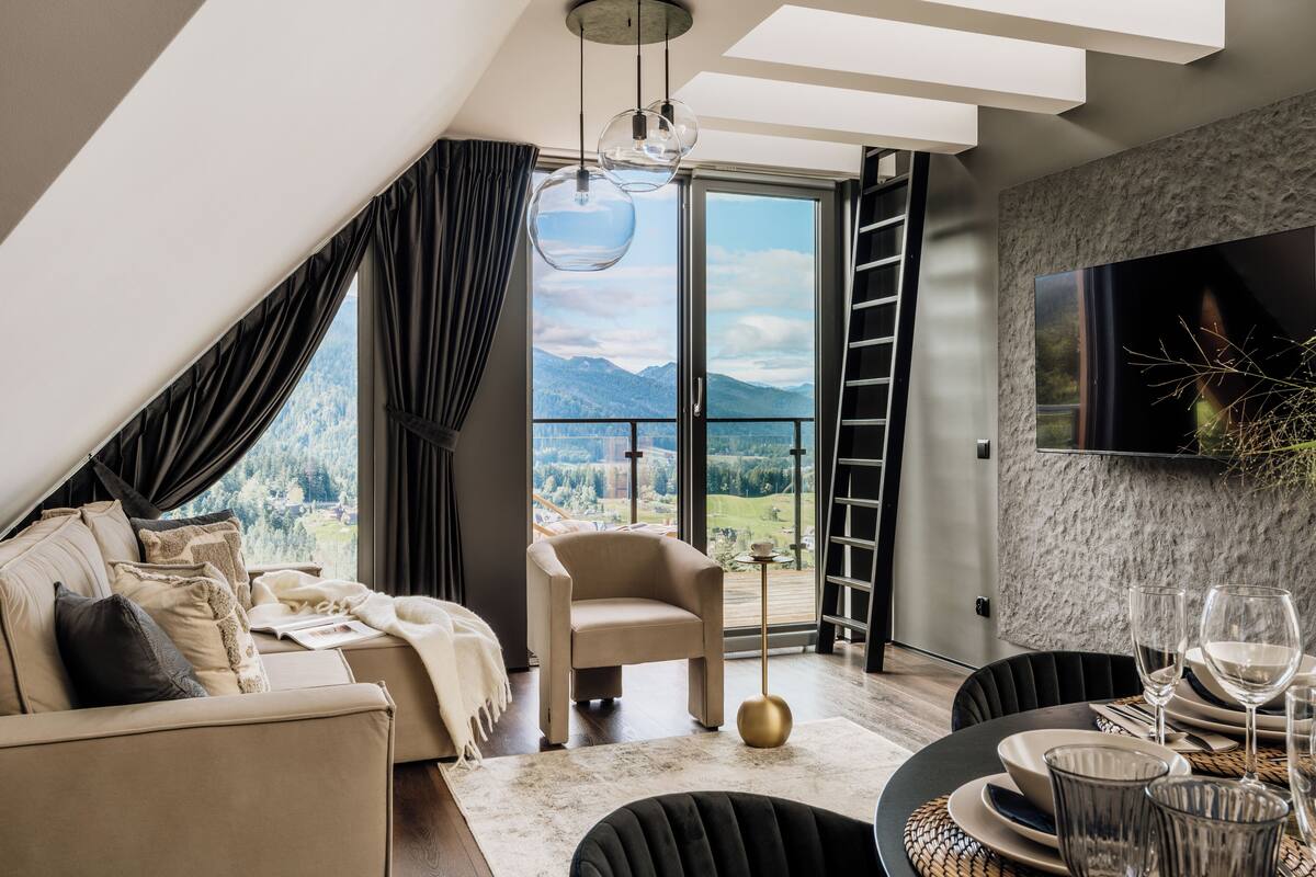 Una sala moderna con sofá, sillón, mesa de comedor y vistas a montañas a través de amplios ventanales.