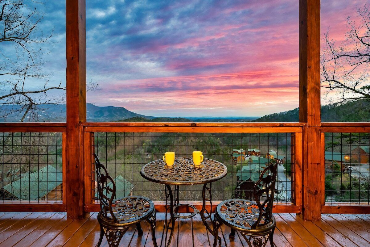 A wooden deck with a table and chairs, a view of a landscape with mountains, and a sunset.