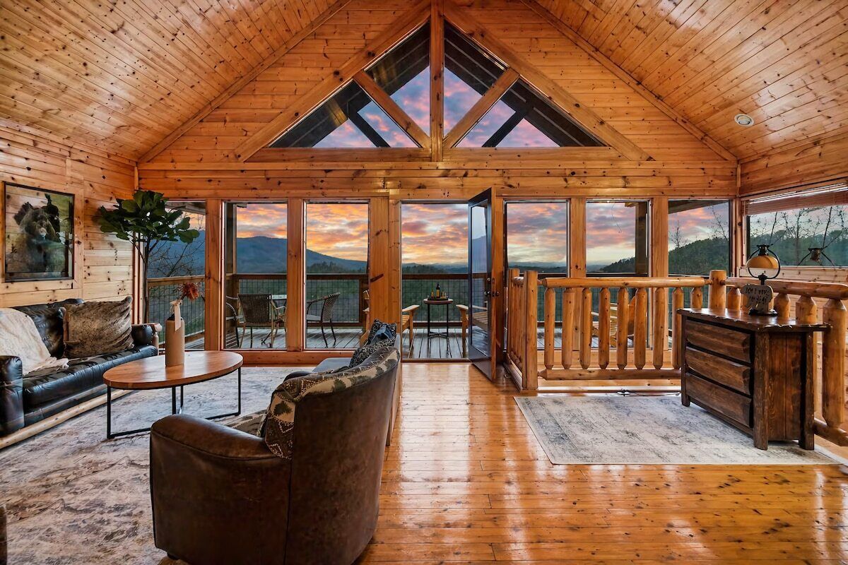 Una cabaña de madera espaciosa con habitación en el altillo, sala de estar con sofá y mesa de centro, y ventanas amplias con vistas a la montaña.