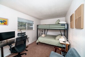 Ein kleines Zimmer mit einem Etagenbett, einem Schreibtisch mit Computer, einem Stuhl und einem Fenster mit Jalousien.