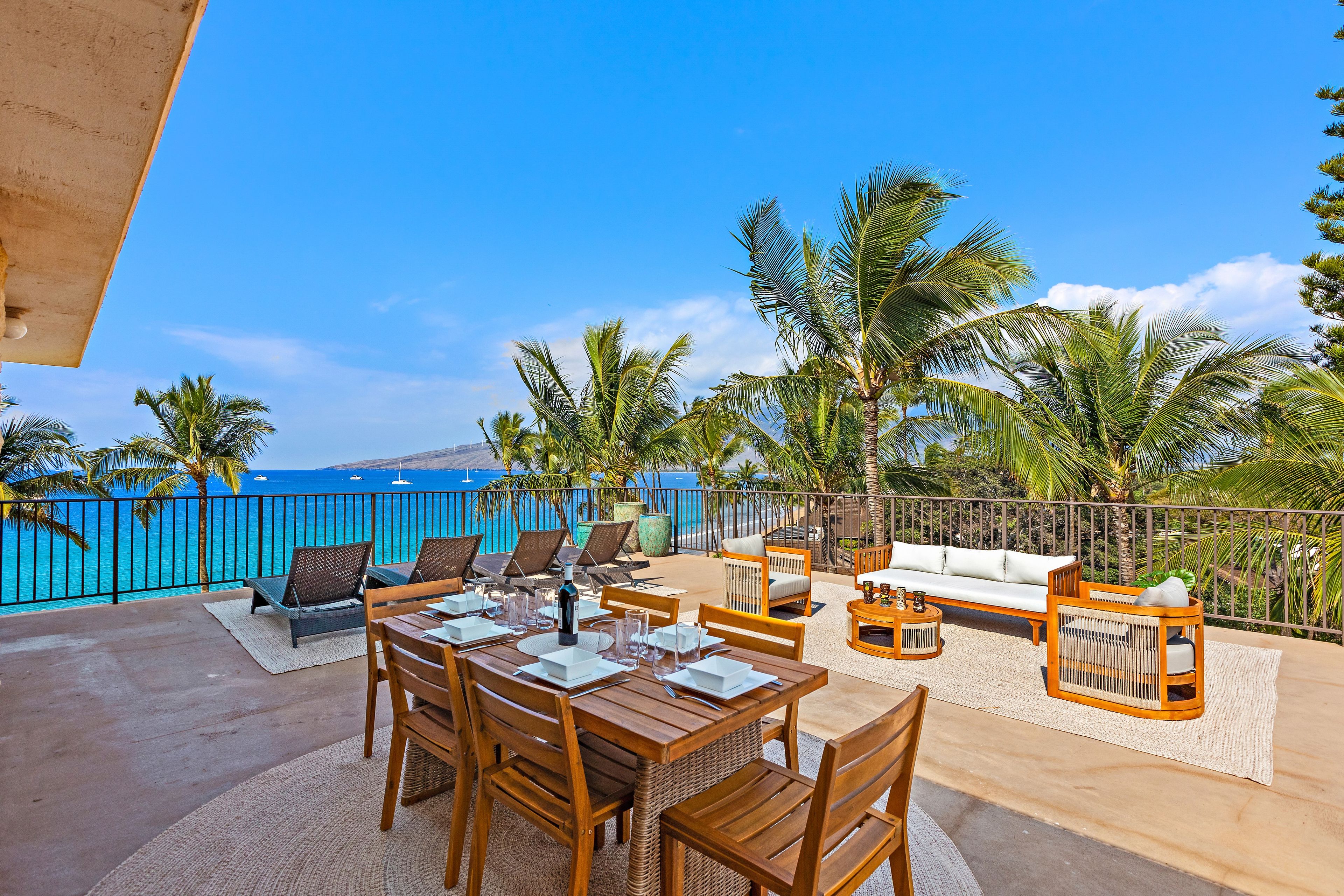 A sunlit outdoor dining area with a view of the ocean, furnished with wooden tables and chairs, and surrounded by palm trees.