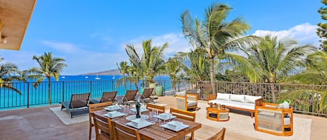 A sunlit outdoor dining area with a view of the ocean, furnished with wooden tables and chairs, and surrounded by palm trees.
