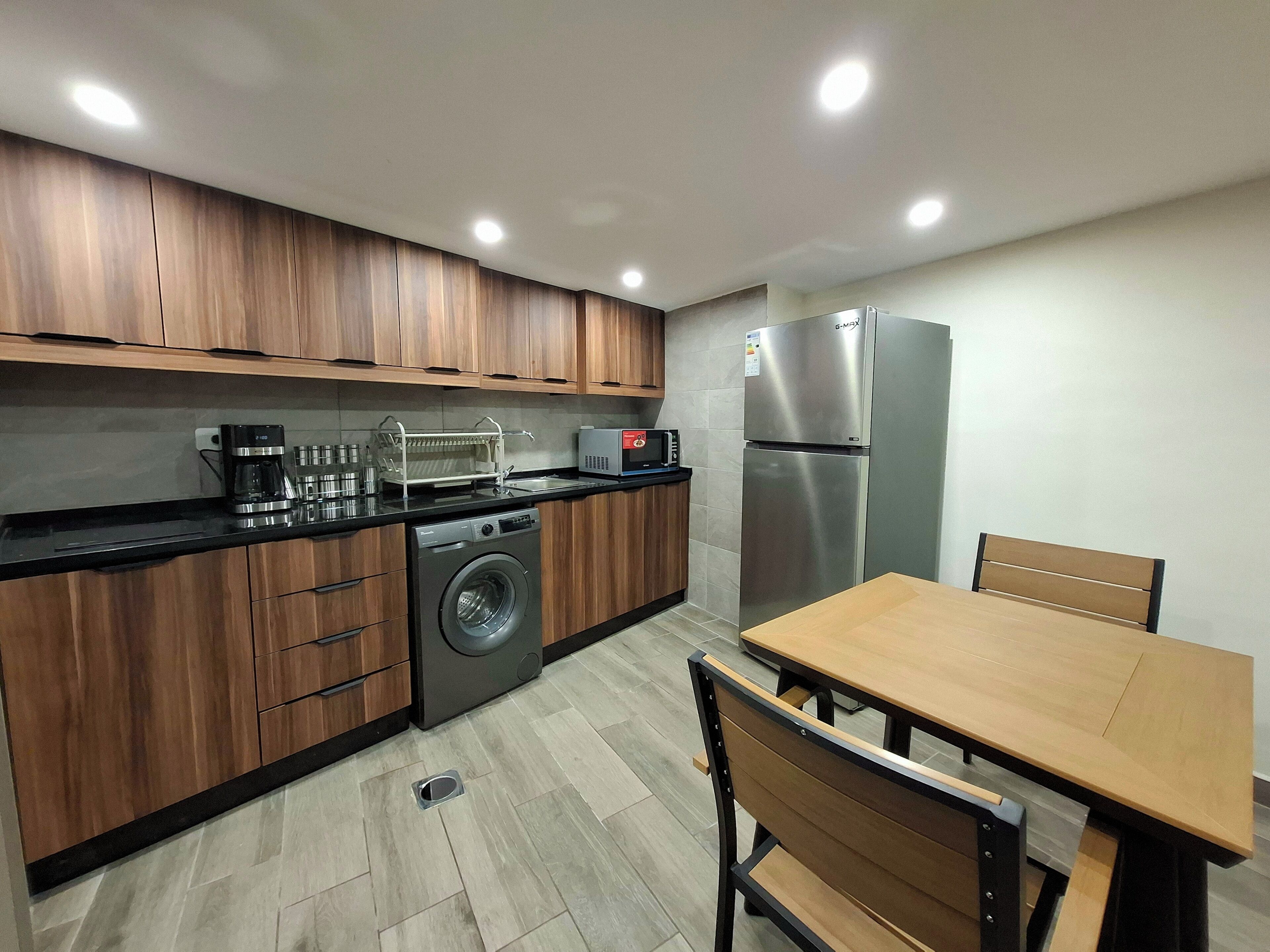 A modern kitchen with wooden cabinets, a washing machine, a refrigerator, a microwave, and a dining table with chairs.