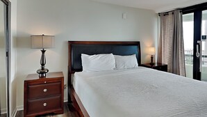 A hotel room with a large bed, a wooden nightstand with a lamp, and a view of the city through the window. - Edgewater #1101 | Gulf Front + Resort Amenities (Miramar Beach)