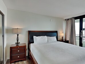 A hotel room with a large bed, a wooden nightstand with a lamp, and a view of the city through the window. - Edgewater #1101 | Gulf Front + Resort Amenities (Miramar Beach)
