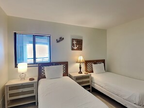 Ein Hotelzimmer mit zwei Einzelbetten, einem Fenster mit Jalousien, einer Nachttischlampe, einem kleinen Tisch und einer Wanddekoration.