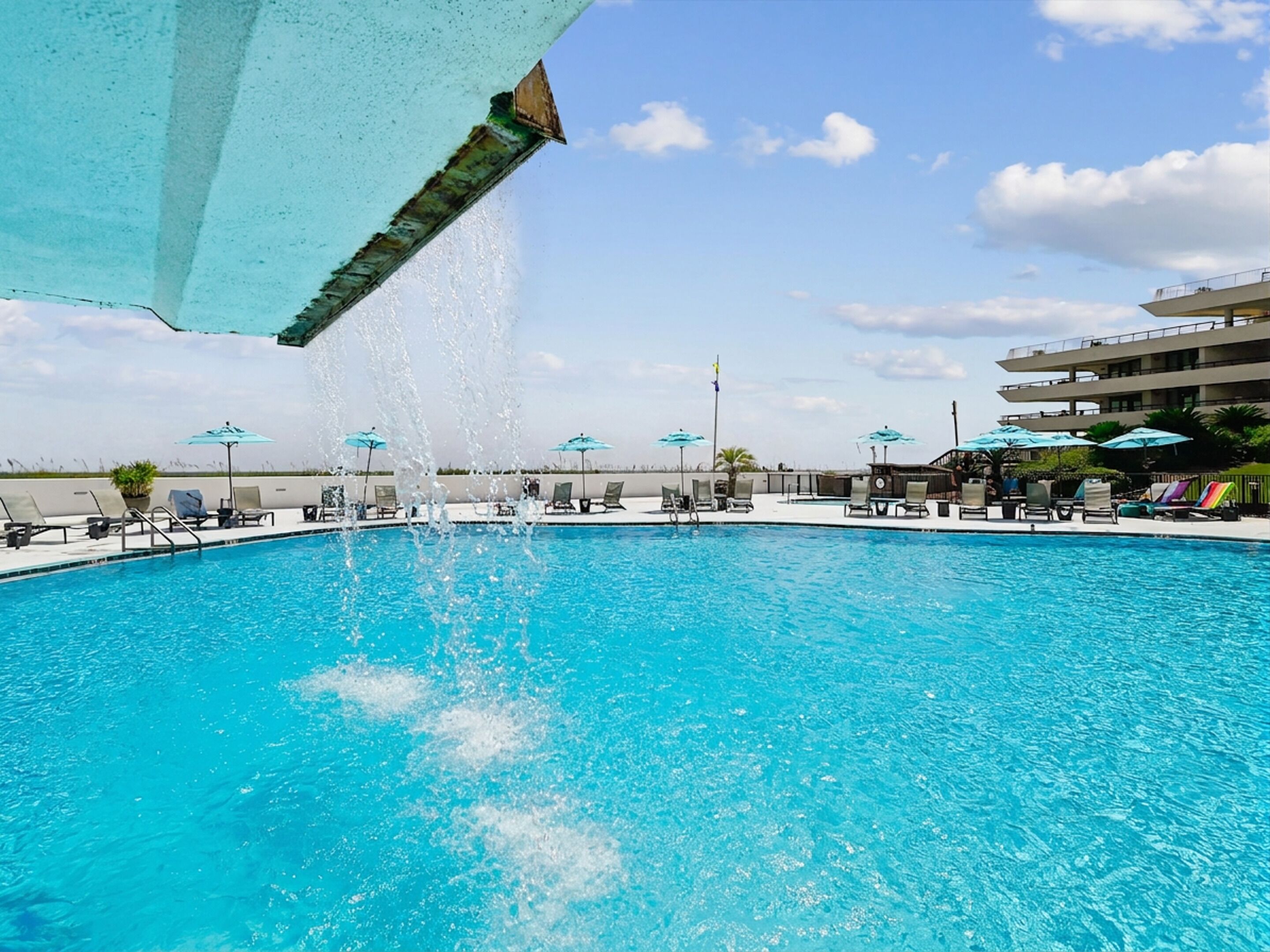 A swimming pool with water jets, lounge chairs, umbrellas, and a building in the background.