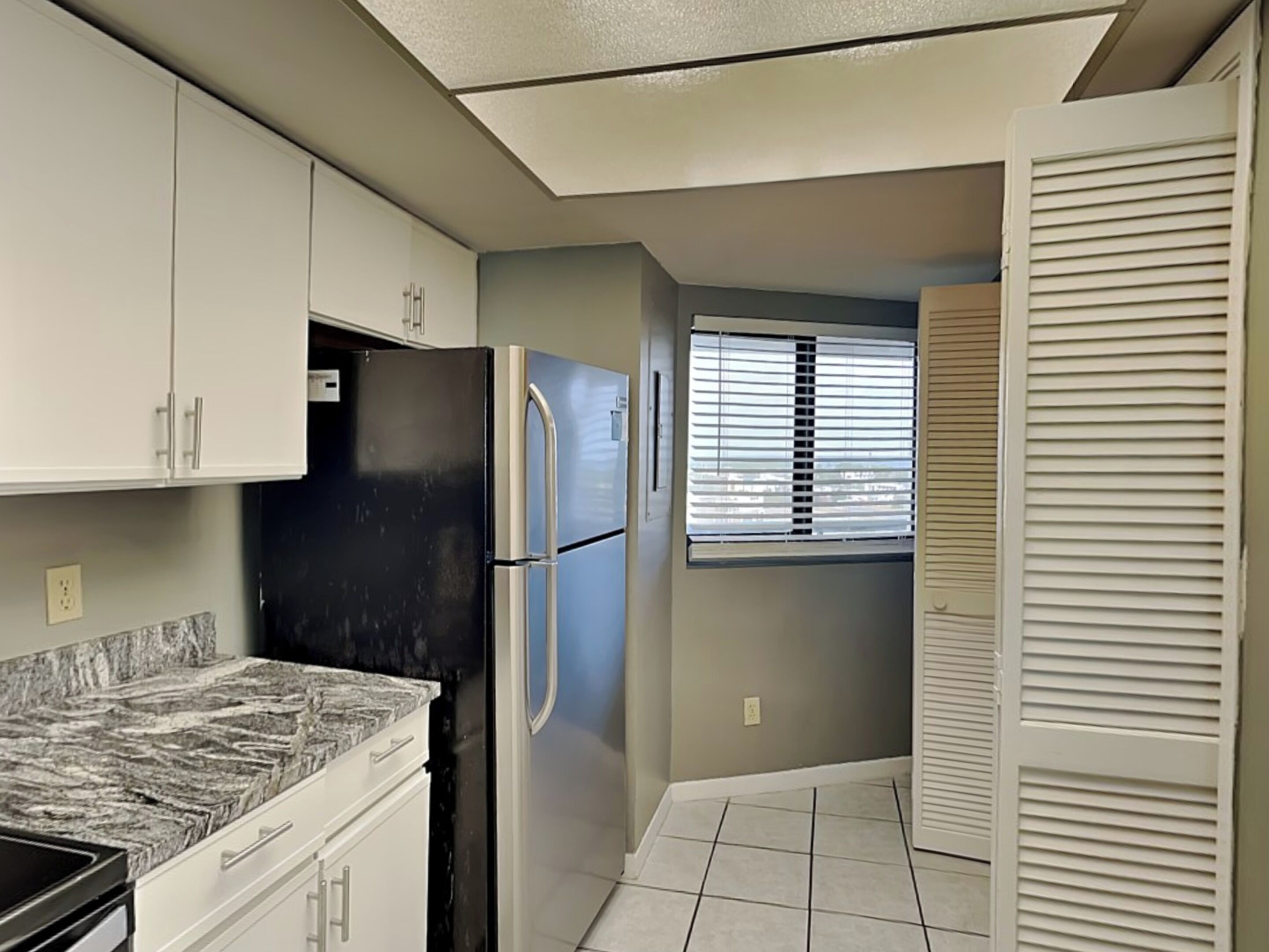 A kitchen with white cabinets, a black refrigerator, a marble countertop, and a window with blinds.