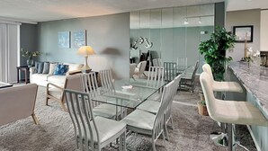 Un salon moderne avec une table à manger en verre, des chaises à revêtement gris, un canapé avec des coussins à motifs, une table d’appoint avec une lampe et un grand miroir.