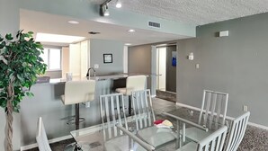 Un espace repas moderne avec une table en verre et des chaises blanches, un coin cuisine avec un bar, et une grande plante en pot.