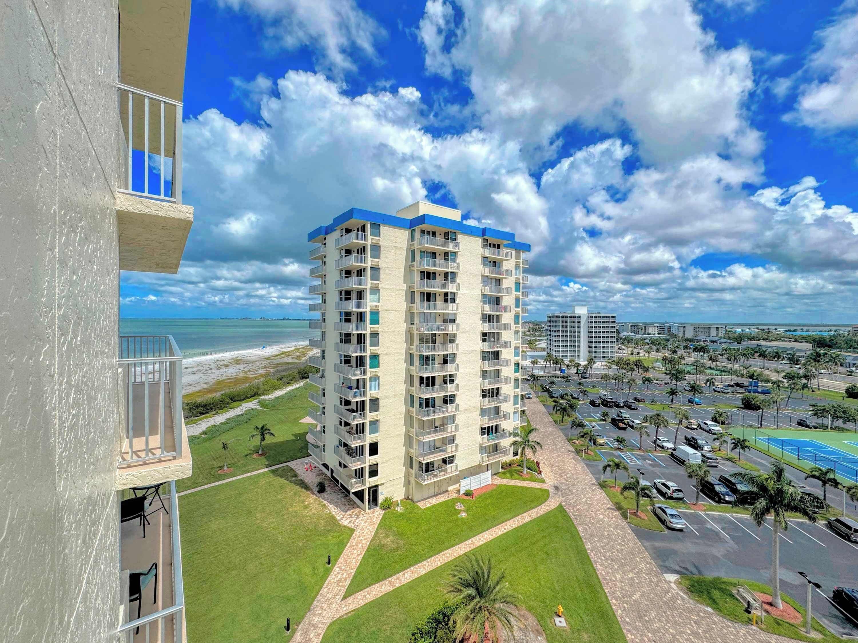 A high-rise building with multiple balconies, a parking lot, and a view of the beach.