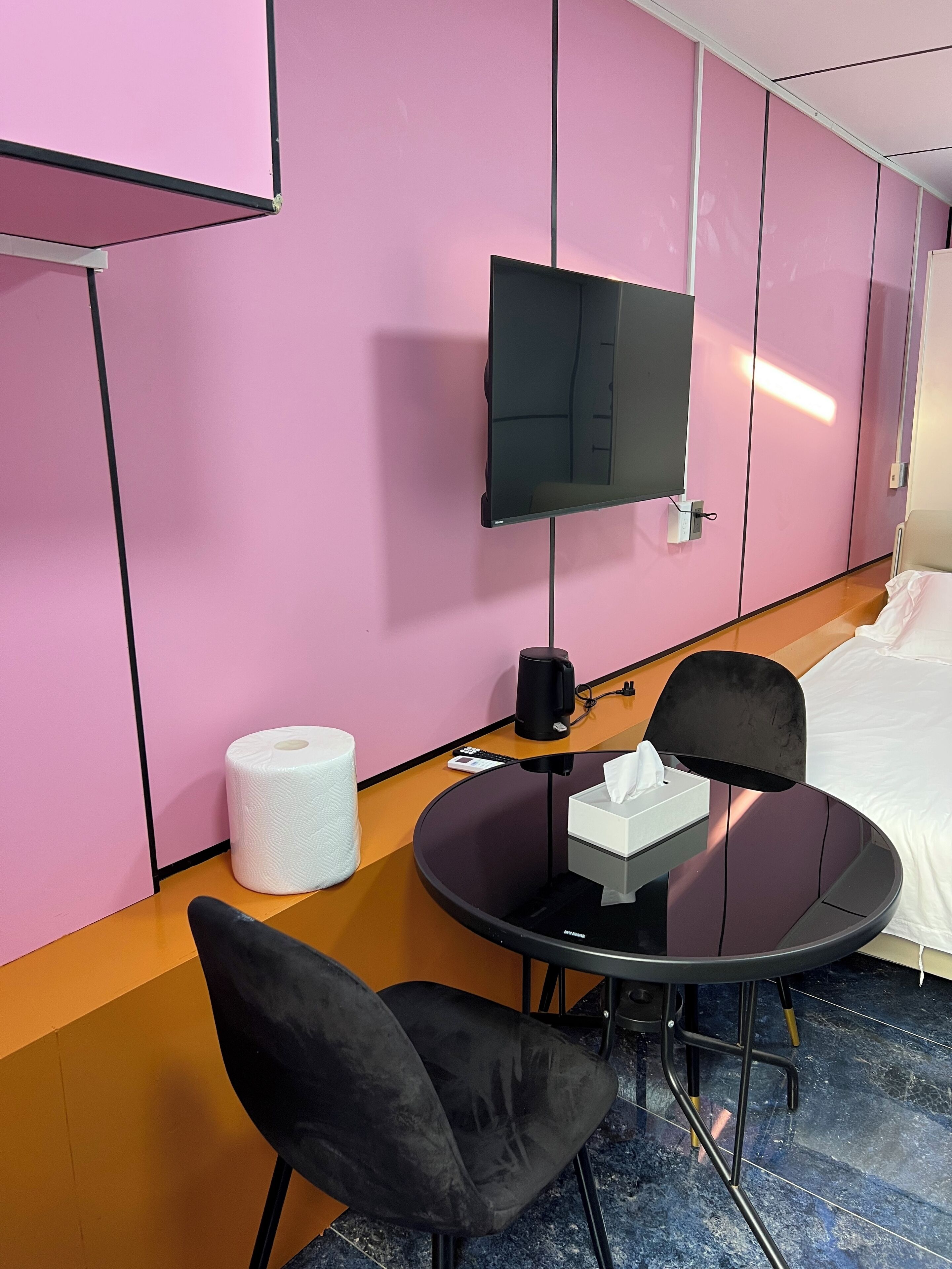 A hotel room with a pink wall, a flat-screen TV mounted on the wall, a round table with a chair, a bed with white bedding, and a roll of toilet paper on the floor.