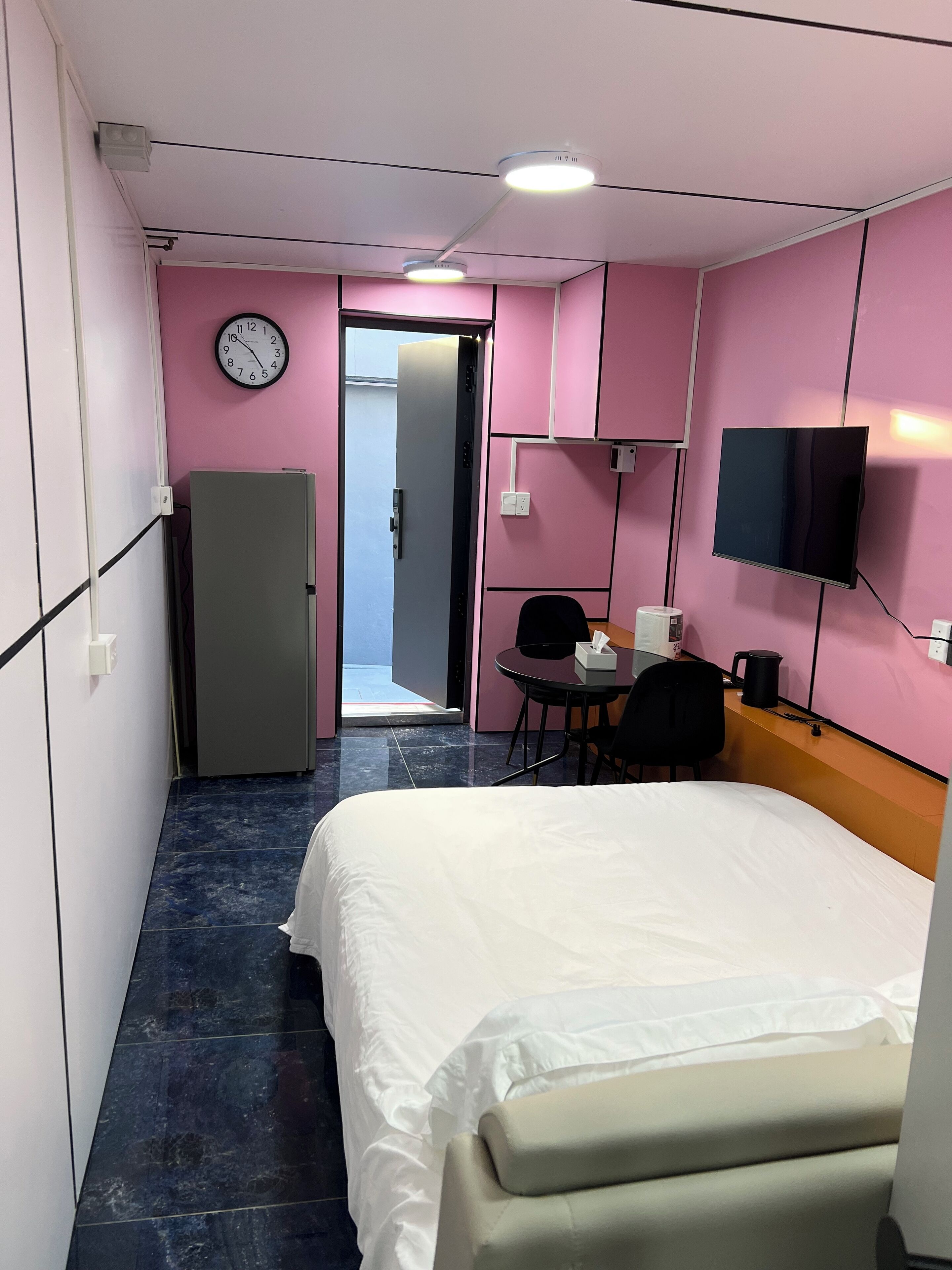 A hotel room with a bed, a desk with a chair, a television mounted on the wall, a refrigerator, and a clock on the wall.
