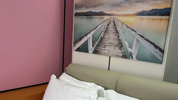 A bed with white linens and pillows, a pink wall, and a painting of a pier extending into a body of water with mountains in the background. - Seafront Studio C4 (Simpson Bay)