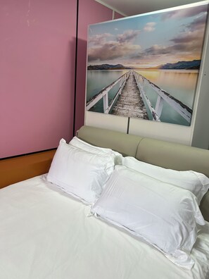 Un lit avec du linge de lit blanc et des oreillers, un mur rose et un tableau représentant une jetée s’avançant dans une étendue d’eau, avec des montagnes en arrière-plan.