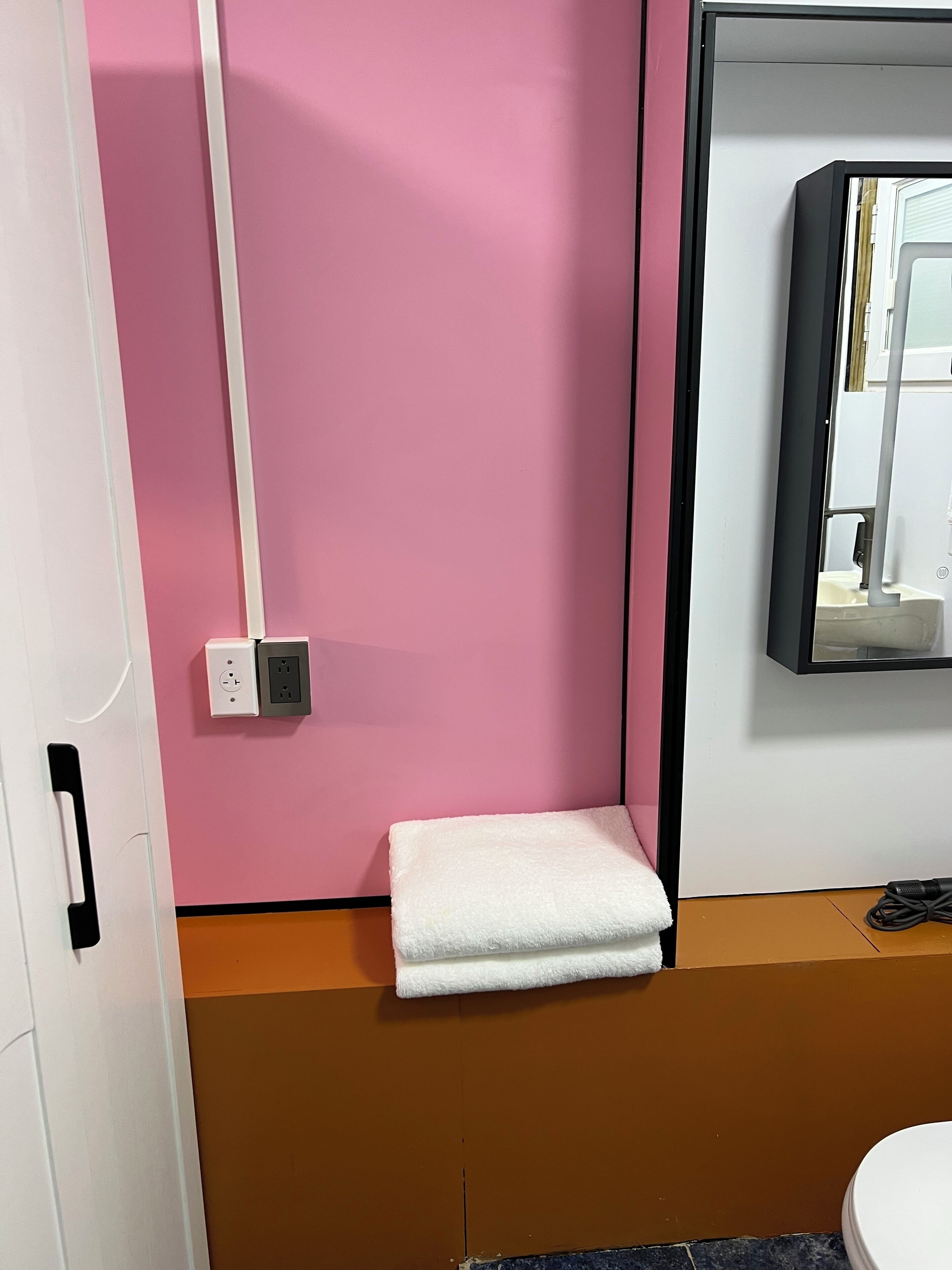 Kylpyhuone, jossa on vaaleanpunainen seinä, valkoinen pyyhe hyllyllä, valkoinen WC-istuin ja peili WC-istuimen yläpuolella.