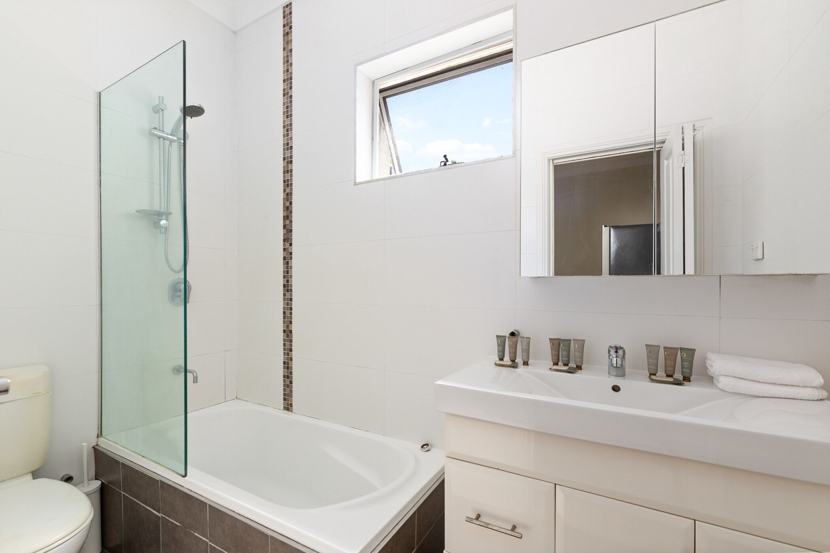 A modern bathroom with a glass-enclosed shower, a bathtub, a vanity with a mirror, and a window providing natural light.