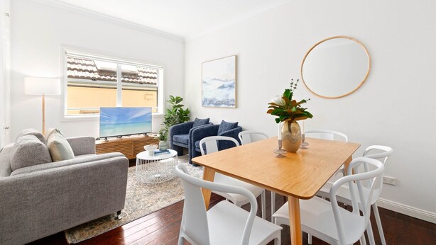 A modern living room with a grey sofa, a wooden dining table with white chairs, a small round table with a lamp, and a large window with a view of the sea. - Bright 3-bed Apartment Near Coogee Beach (Sydney)