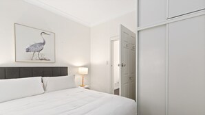 A neatly made bed with white linens and a gray headboard, a bedside lamp, and a framed bird artwork on the wall. - Bright 3-bed Apartment Near Coogee Beach (Sydney)