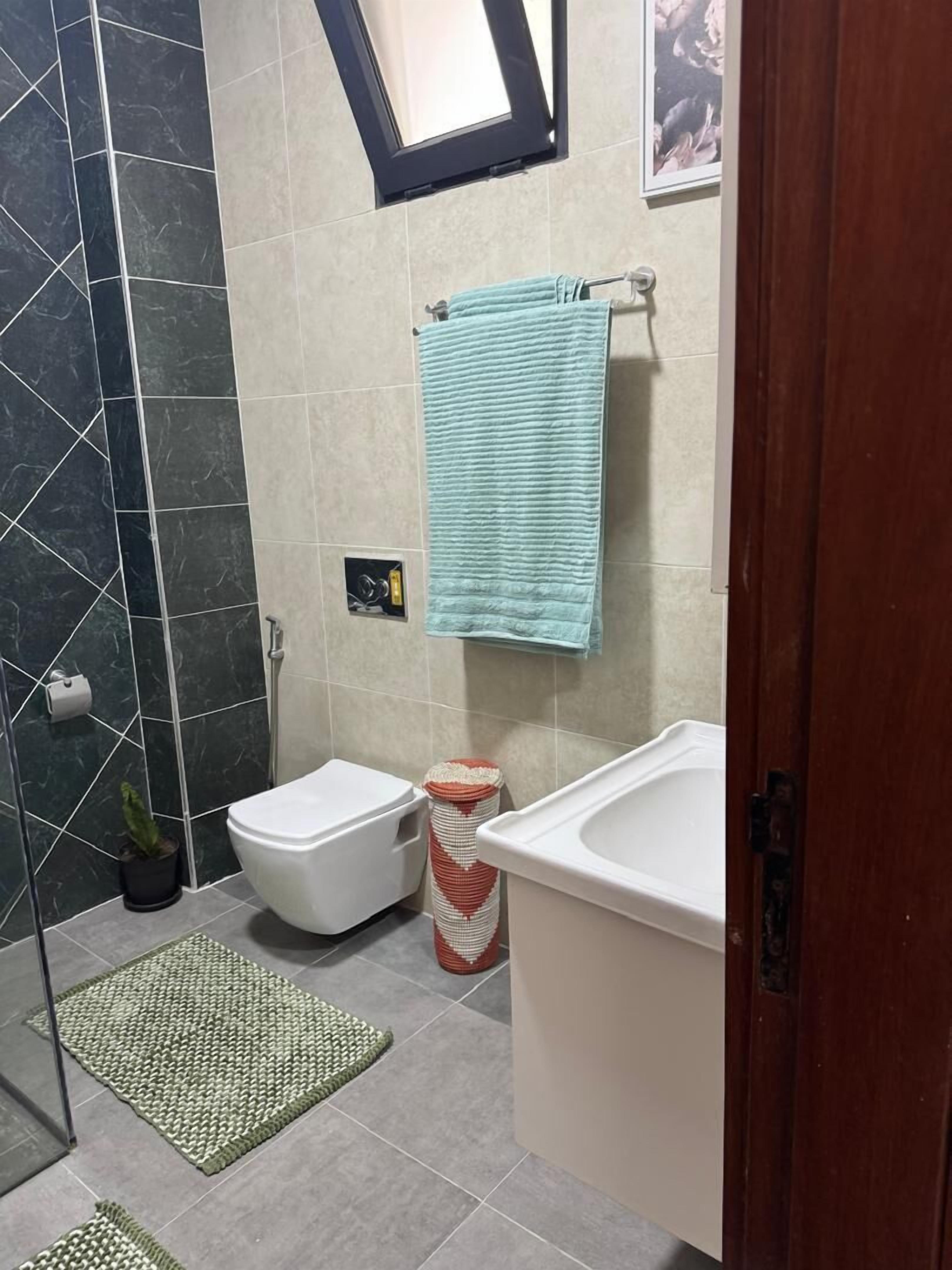 Kylpyhuoneessa on wc, bidee ja kylpyamme. Siellä on myös pyyheteline, jossa on pyyhe, pieni kasvi ja vihreä matto lattialla.