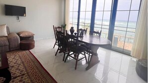 A spacious living room with a large dining table and chairs, a flat-screen TV mounted on the wall, and a large window offering a view of the ocean. - Residence Al Awane (Dakar)
