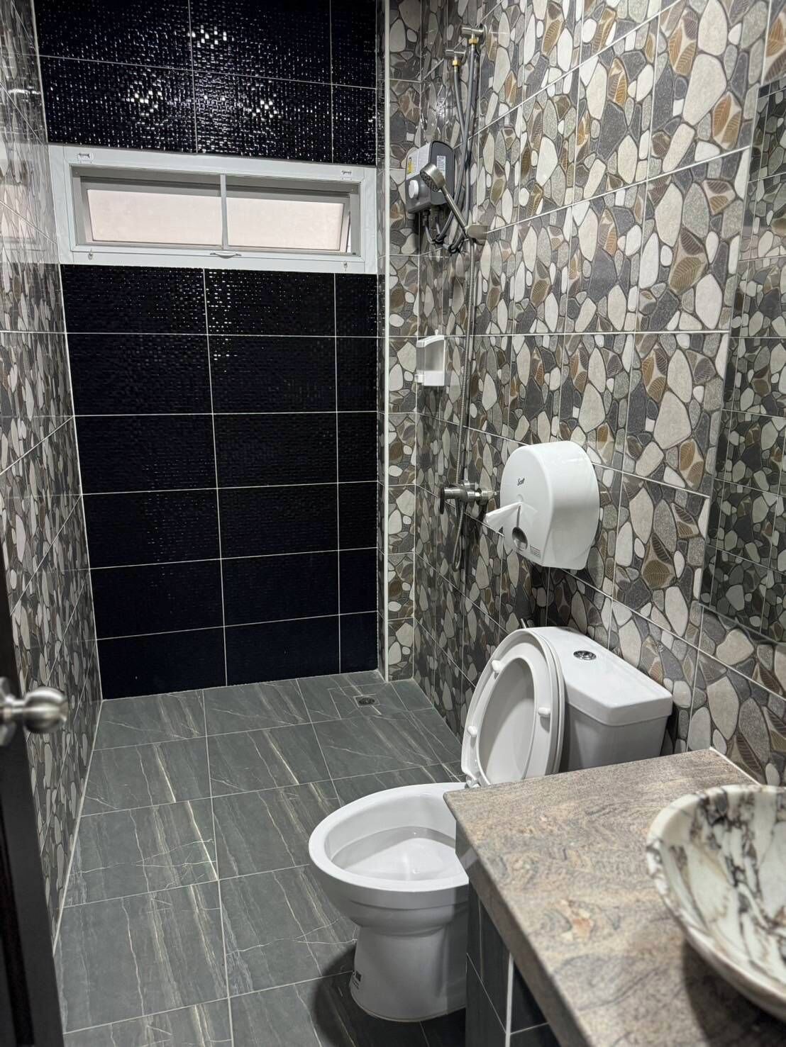 A bathroom with a toilet, bidet, and a hand dryer. The walls are tiled with a mix of black and multicolored stone tiles.