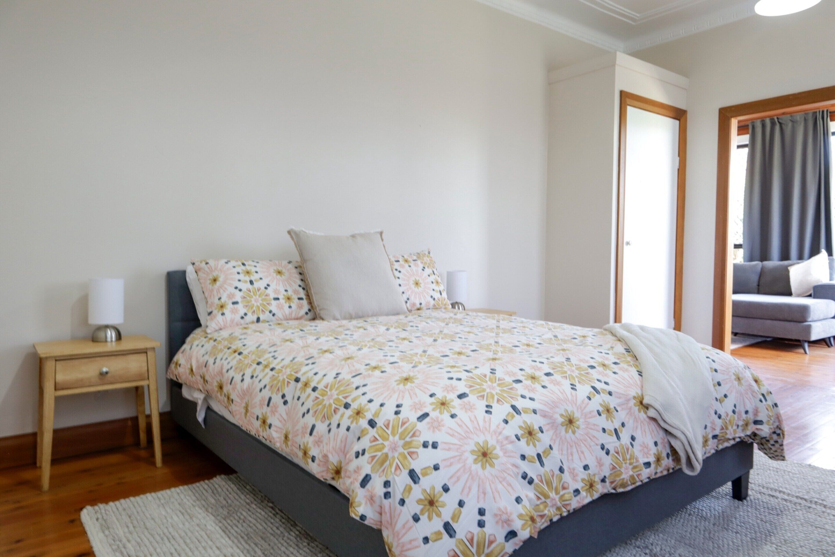 Une chambre à coucher avec un lit, une table de chevet, une lampe, un canapé gris et une fenêtre avec des rideaux.