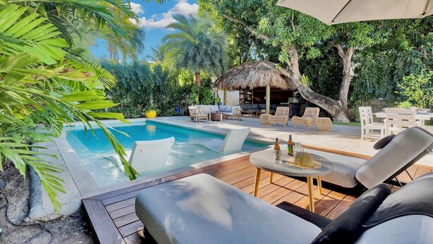 Un área junto a la piscina con sillones de descanso, una mesa con bebidas y una estructura de techo de paja.
