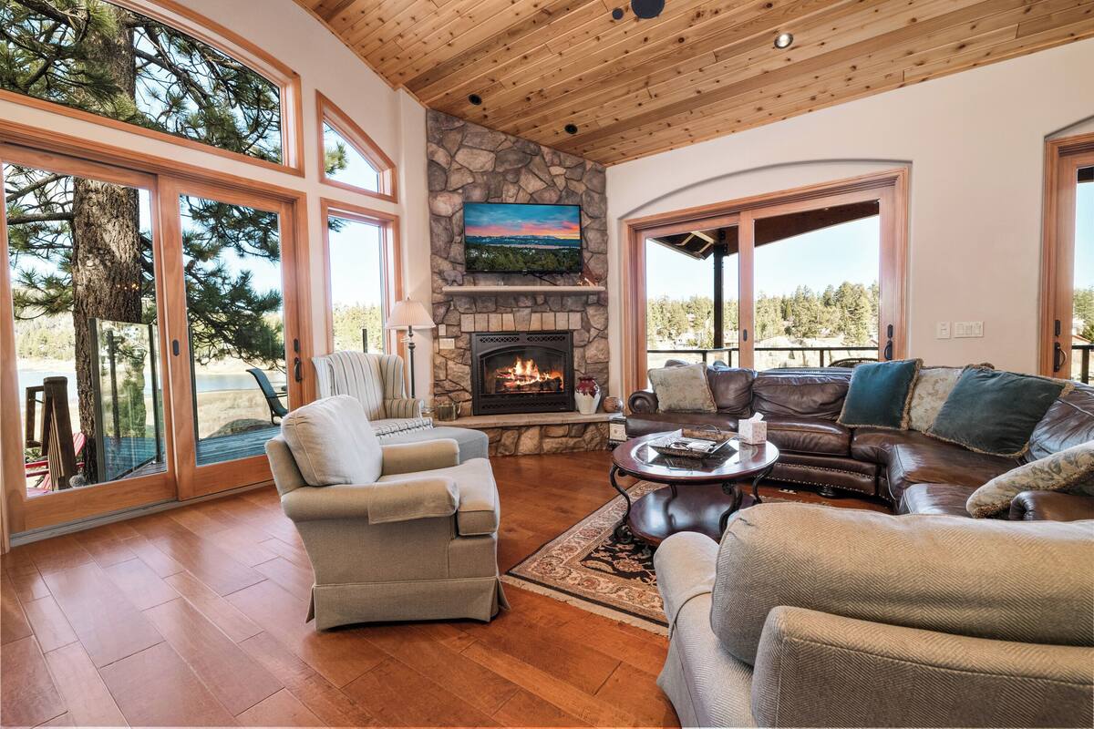 A spacious living room with a stone fireplace, leather sofas, and large windows offering a view of trees and a distant landscape.