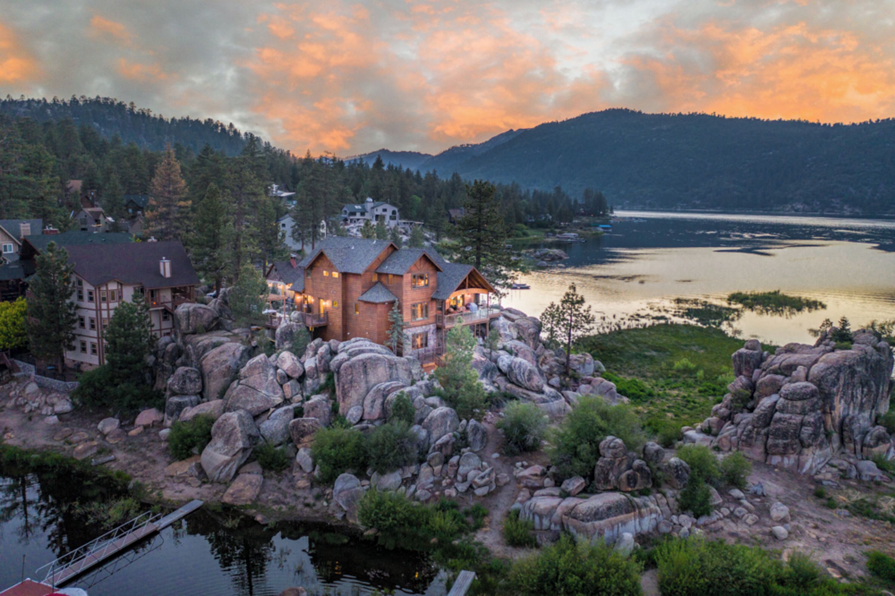 A lakeside lodge with multiple buildings, surrounded by large rocks and trees, overlooking a calm lake and forested hills.