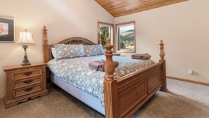 A bedroom with a wooden bed, a nightstand with a lamp, and a window with a mountain view. - Boulder Cove Lakefront Estate (Big Bear Lake)