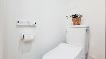 Un baño pequeño y moderno con inodoro blanco, pared blanca, suelo blanco, techo blanco, tanque de descarga empotrado blanco, dispensador de papel higiénico blanco y una pequeña planta en el tanque del inodoro.