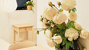 Een kamer met een houten tafel, een vaas witte bloemen, een klein houten krukje en een televisie aan de muur.