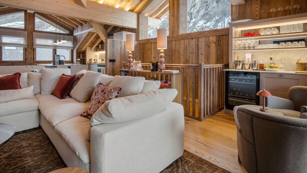 Ein gemütliches Wohnzimmer mit einer Couch und Sesseln, Holzbalken, einer Küchenzeile und einer Bar.