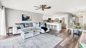 Ein modernes Wohnzimmer mit einer Couch, einem Couchtisch und einem Deckenventilator. Es gibt eine Küchenzeile mit weißen Schränken und einem Esstisch.