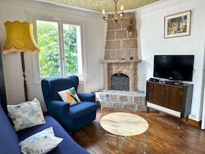 A living room with a blue sofa, a round coffee table, a stone fireplace, a television on a wooden cabinet, and a lamp. - House with enclosed garden in Ploumanac’h (Perros-Guirec)