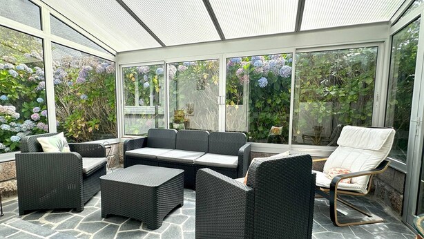 Ein überdachter Patio-Bereich mit Korbmöbeln, Steinböden und großen Fenstern mit Blick auf einen Garten mit Hortensien.