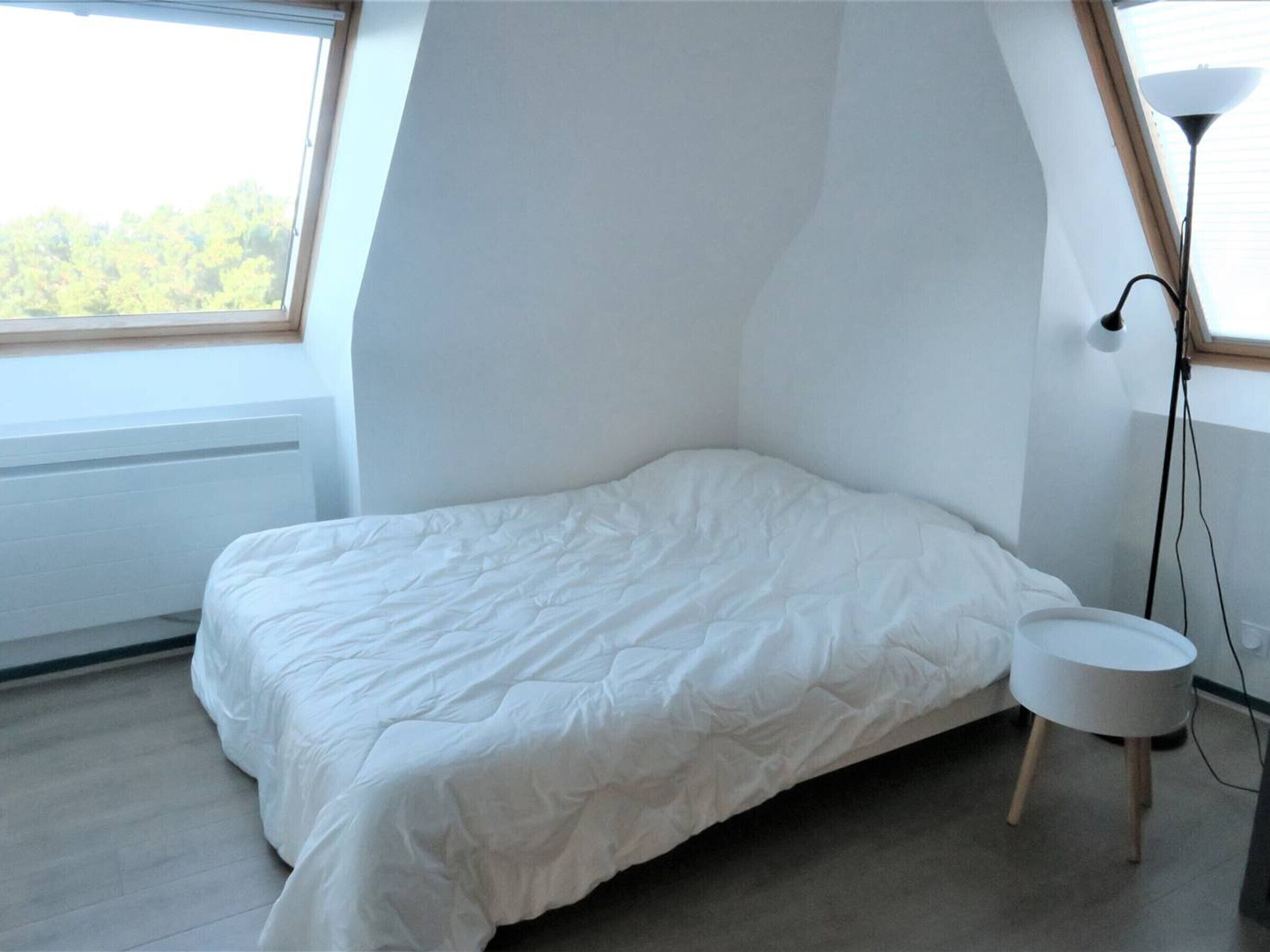 Un dormitorio con una cama, una mesita de noche redonda y blanca, una lámpara y un ventanal con vistas a árboles.