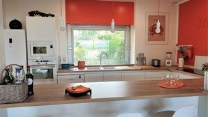 A modern kitchen with a central island, white cabinets, a red accent wall, and a window providing a view of greenery outside. - Beachfront house in Trébeurden with garden and terrace (Trébeurden)