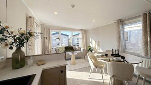 Una sala moderna con sofá, mesa de comedor y sillas, zona de cocina con mostrador y armarios, y ventanas amplias con cortinas.