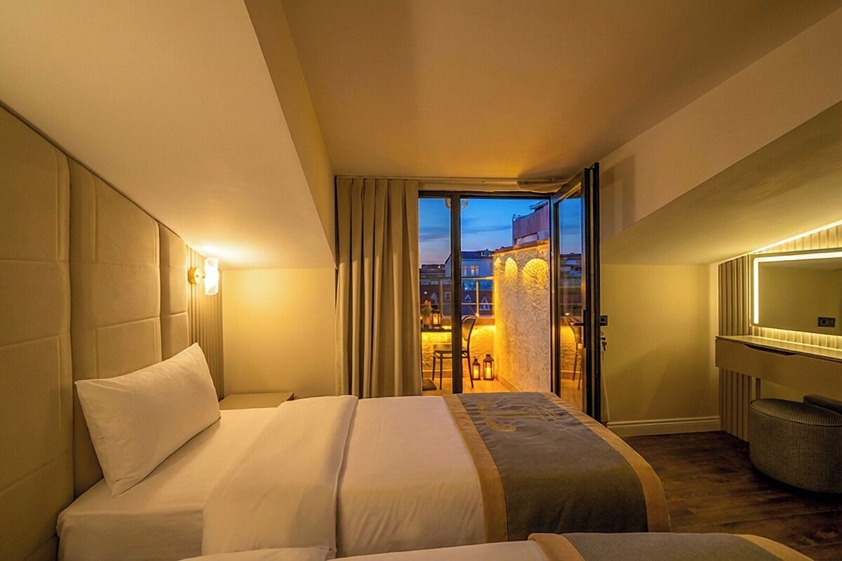 A hotel room with a large bed, a desk, a chair, a mirror, and a view of the city at dusk.