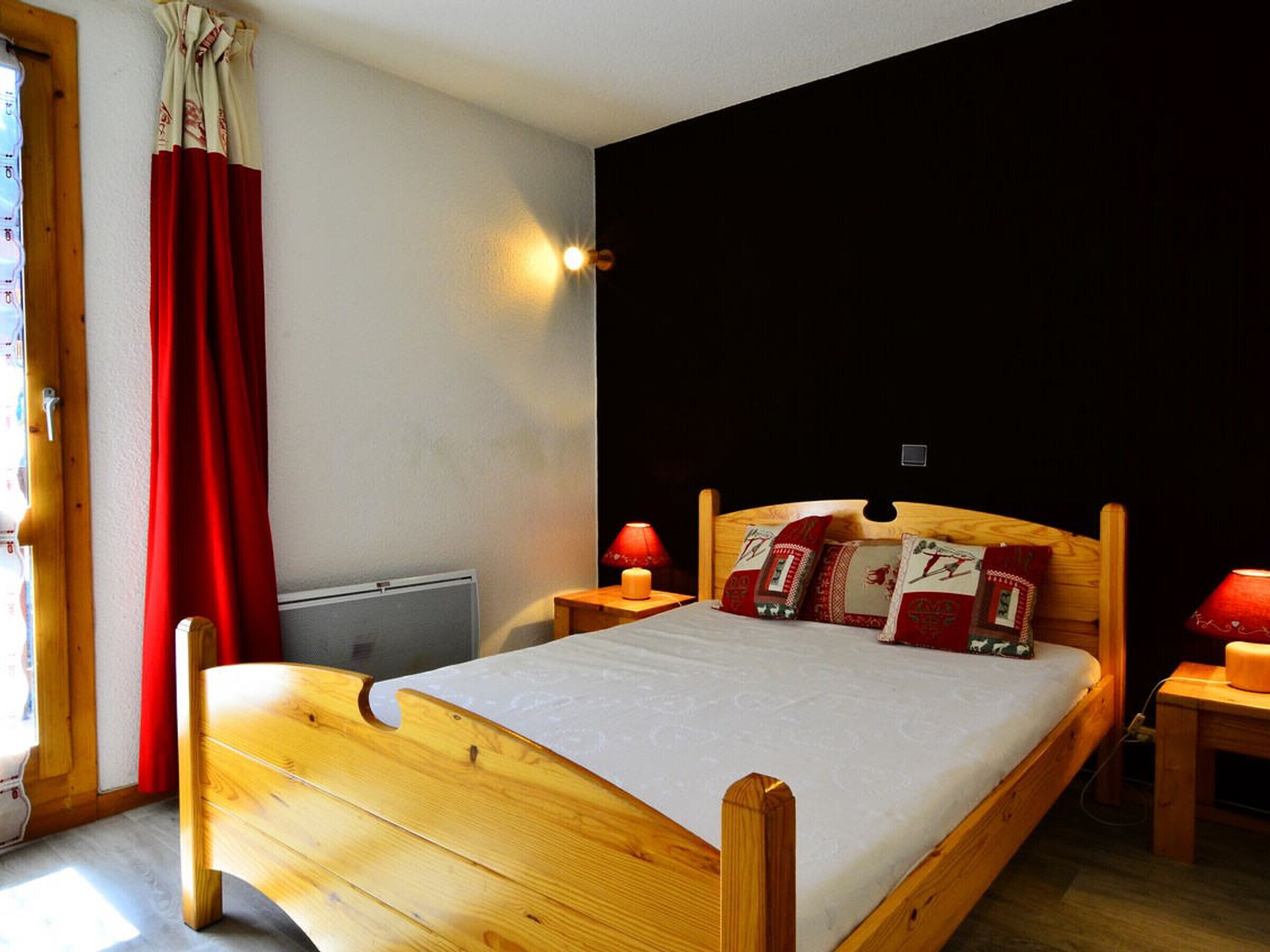 Una cama de madera con colchón blanco, almohadas rojas y beige, y una mesita de noche con una lámpara roja.