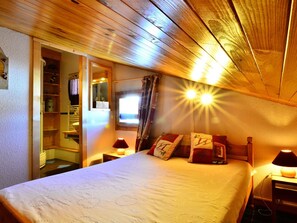 Ein gemütliches Schlafzimmer mit Holzbalkendecke, einem Bett mit gemusterten Textilien, zwei Nachttischlampen und einem Fenster mit Vorhängen.