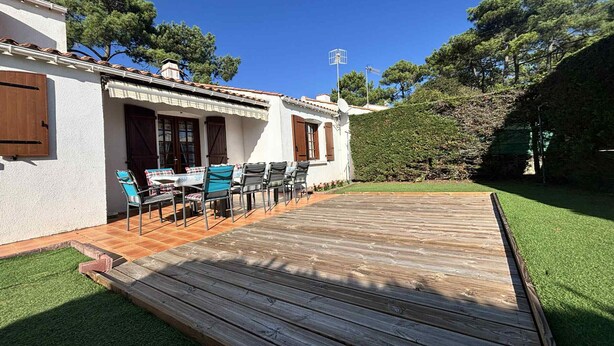 Un patio con mobiliario exterior, una terraza de madera y un edificio blanco con contraventanas marrones.