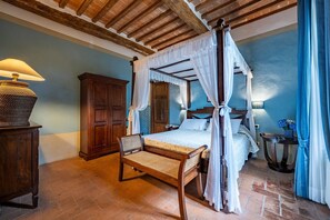 A four-poster canopy bed with a wooden headboard, a wooden nightstand with a lamp, and a wicker chair. - Podere Sant'Eugenio 14, Emma Villas (Chiusdino)