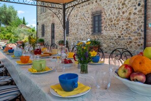 An outdoor dining area with a stone wall backdrop, a long table set with various dishes, cups, and a fruit bowl. - Podere Sant'Eugenio 14, Emma Villas (Chiusdino)